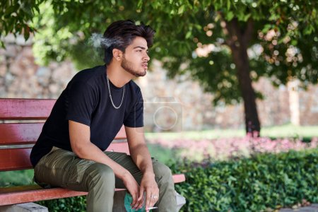 Photo for Attractive indian man smoker exhales cigarette smoke portrait in black t shirt and silver neck chain sitting on bench in public park, hindu male smoking close up portrait. Handsome indian man portrait - Royalty Free Image