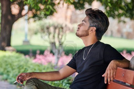 Photo for Attractive indian man smoker exhales cigarette smoke portrait in black t shirt and silver neck chain sitting on bench in public park, hindu male smoking close up portrait. Handsome indian man portrait - Royalty Free Image