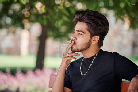 Photo for Young indian man smoker portrait in black t shirt and silver neck chain sitting on bench in public park, hindu male smoking close up portrait. Handsome indian man portrait with thick hair in city park - Royalty Free Image
