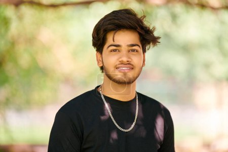 Smiling young indian man candid portrait in black t shirt and silver neck chain on outdoor public park background, close up hindu attractive male portrait. Handsome indian man portrait with thick hair