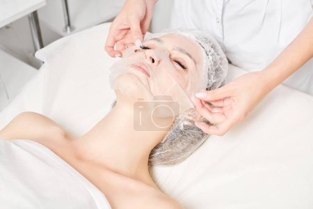 Téléchargez les photos : Esthéticienne lisse masque de feuille sur le visage de la femme pour réhydrater la peau du visage, procédure cosmétique anti rides dans le salon de beauté spa. Cosmetologue appliquant un masque hydratant sur le visage féminin - en image libre de droit