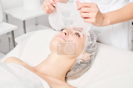 Téléchargez les photos : Esthéticienne enlever masque de feuille sur le visage de la femme pour réhydrater la peau du visage, procédure cosmétique anti-âge dans le salon de beauté spa. Cosmetologue enlever masque de feuille hydratante sur le visage féminin - en image libre de droit