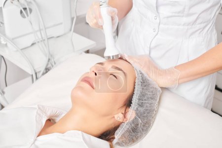 Cosmetologist makes ultrasound skin tightening for rejuvenation woman face using phonophoresis, anti aging cosmetic procedure with in beauty spa salon. Beautician makes ultrasonic skincare lifting