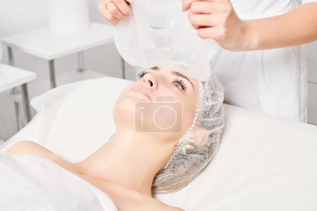 Téléchargez les photos : Esthéticienne enlever masque de feuille sur le visage de la femme pour réhydrater la peau du visage, procédure cosmétique anti-âge dans le salon de beauté spa. Cosmetologue enlever masque de feuille hydratante sur le visage féminin - en image libre de droit