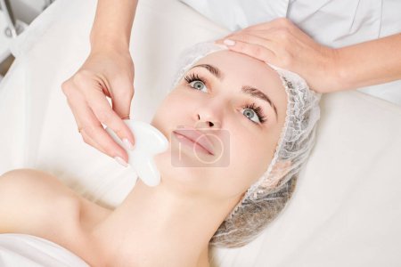 Photo for Cosmetologist making facial massage with Gua Sha stone of woman face skin for lymphatic drainage, anti aging skincare cosmetic procedure in beauty spa salon. Face massage with Gua Sha tool - Royalty Free Image