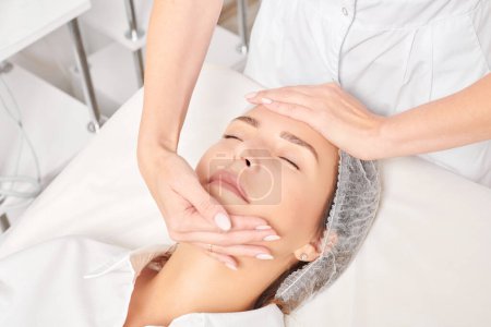 Photo for Cosmetologist massages woman face skin after rubbing moisturizing cream for rejuvenation, anti aging cosmetic procedure in beauty spa salon. Beautician hands massages female face for relax - Royalty Free Image