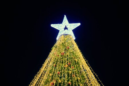 Photo for Christmas tree with big white star topper decorated yellow garlands and colorful decorative bulbs on night blue sky background, outdoor holiday atmosphere. Festive Christmas tree with yellow lights - Royalty Free Image