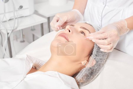 Cosmetologue applique des capsules d'huile faciale sur le visage de la femme pour le rajeunissement, procédure cosmétique anti-âge dans le salon de beauté spa. Les mains d'esthéticienne dans les gants tiennent des capsules d'huile pour se nourrir