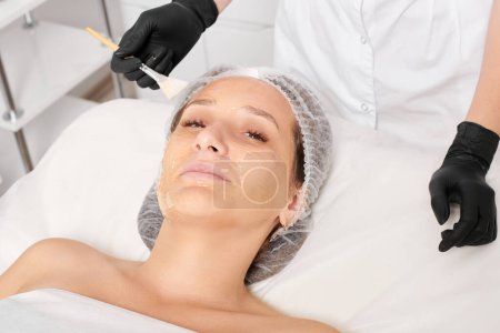 Photo for Beautician applies honey mask on woman face for moisturize face skin, anti aging cosmetic procedure in beauty spa salon. Cosmetologist in black gloves holds cosmetic brush for applying honey mask - Royalty Free Image