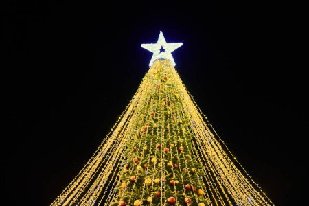 Photo for Christmas tree with yellow garlands, decorative bulbs and big white star topper at night blue sky background, outdoor holiday atmosphere. Festive Christmas tree with luminous yellow light garlands - Royalty Free Image