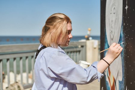 Young female painter passionately paints picture with paintbrush for outdoor street exhibition, female artist engrossed in creating vibrant artwork at bright sunny day