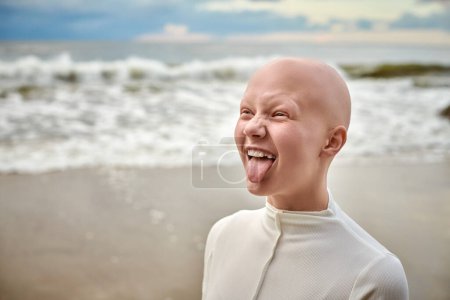 Photo for Hairless girl with alopecia make faces in white futuristic costume on sea background, close up portrait of bald pretty teenage girl showcasing unique beauty and identity with pride, unusual alien girl - Royalty Free Image