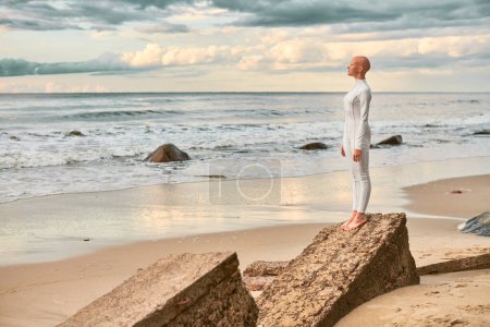 Ganztägiges Porträt eines jungen haarlosen Mädchens mit Alopezie im weißen futuristischen Anzug, das am steinernen Strand am Meer steht, metaphorische surreale Szene mit glatzköpfigem Teenager-Mädchen strahlt Zuversicht und einzigartige Schönheit aus