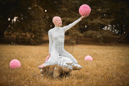 Junges haarloses Mädchen mit Alopezie in weißem Tuch sitzt auf teergrader Figur und hält rosa Ball in der Hand auf Herbst-Rasenpark, surreale Szene mit glatzköpfigem Teenager-Mädchen beschäftigt sich mit symbolischen Elementen