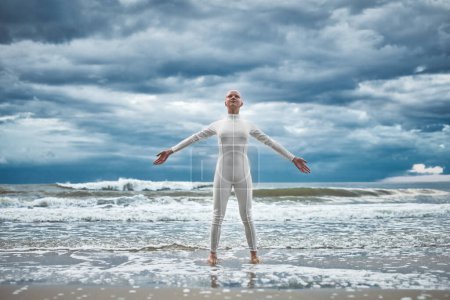 Photo for Happy hairless girl with alopecia in white futuristic suit stands with spread arms on beach bathed by ocean waves, performance of bald strong female artist symbolizing self acceptance - Royalty Free Image