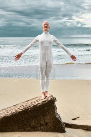 Young hairless girl with alopecia in white futuristic suit standing on sea beach spread her hands, metaphoric surreal performance with bald pretty teenage girl exudes confidence and unique beauty