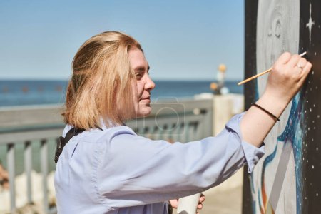 Junge Malerin malt leidenschaftlich Bild mit Pinsel für Straßenausstellung im Freien, weibliche Künstlerin vertieft in die Schaffung lebendiger Kunstwerke bei strahlendem Sonnenschein