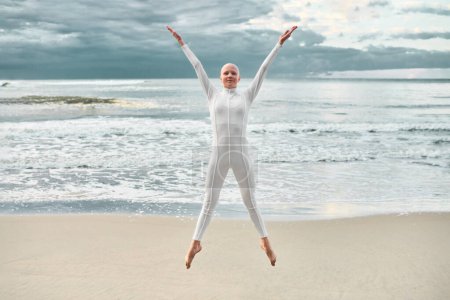Joyful hairless girl with alopecia in white futuristic suit jumping spreading arms and legs apart on sea sandy beach, metaphoric performance with bald teenage girl exudes confidence and happiness