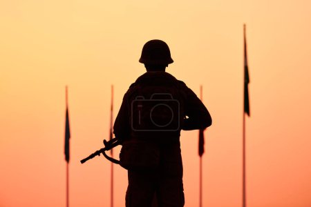 Rückansicht der Silhouette des Soldaten in militärischer Ausrüstung hält Waffe gegen blutigen Sonnenuntergang und gesenkten Flaggen der Länder, schwere Szene symbolisiert feierliche Ehre und Opfer im Namen des Friedens