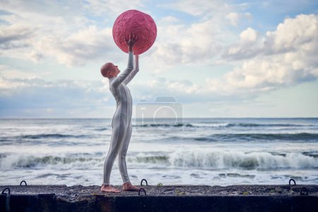 Junges haarloses Mädchen mit Alopezie im weißen futuristischen Anzug, der auf einem Betonzaun steht und eine rosafarbene Kugel auf Meeresgrund hält, metaphorische surreale Performance mit einem glatzköpfigen hübschen Teenager-Mädchen