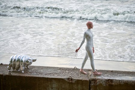 Junges haarloses Mädchen mit Alopezie im weißen futuristischen Anzug, das auf einem Betonzaun mit Spielzeug auf Meeresgrund spaziert, glatzköpfige hübsche Mädchen symbolisiert Mut und Akzeptanz der einzigartigen Erscheinung