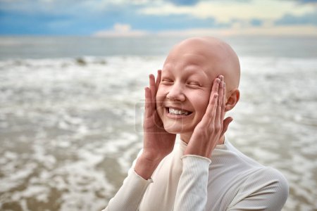 Photo for Hairless girl with alopecia make faces in white futuristic costume on sea background, close up portrait of bald pretty teenage girl showcasing unique beauty and identity with pride, unusual alien girl - Royalty Free Image