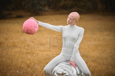 Joven chica sin pelo con alopecia en tela blanca se sienta en la figura tardía y sostiene la bola rosa en la mano en el parque de césped de otoño, escena surrealista con chica adolescente calva se involucra con elementos simbólicos
