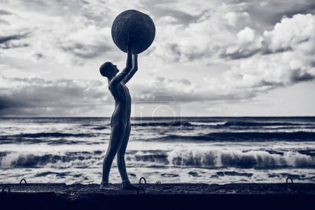 Jeune fille sans cheveux avec alopécie en costume futuriste blanc debout sur une clôture en béton et tenant la sphère rose sur fond de mer, bleu foncé tonique, scène surréaliste métaphorique avec adolescente chauve