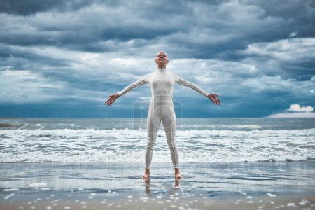 Happy hairless girl with alopecia in white futuristic suit stands with spread arms on beach bathed by ocean waves, performance of bald strong female artist symbolizing self acceptance