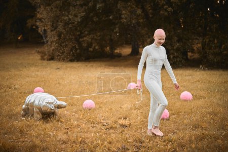Porträt eines jungen haarlosen Mädchens mit Alopezie in weißem Tuch zu Fuß tardigrade Spielzeug im Herbst Park, surreale Szene mit glatzköpfigen Teenager-Mädchen reflektieren über verflochtene Fäden des Lebens und der Kunst