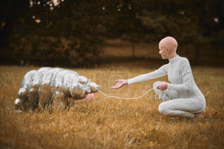 Portrait of young hairless girl with alopecia in white cloth playing with tardigrade toy in fall park, surreal scene with bald teenage girl reflect on intertwining threads of life and art