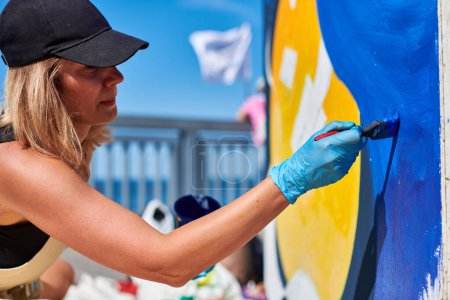 Erwachsene Malerin in schwarzer Mütze malt leidenschaftlich Bild mit Pinsel für Straßenausstellung im Freien mit lebendigen Farben, visuelles Spektakel durch ihre ausdrucksstarken Pinselstriche