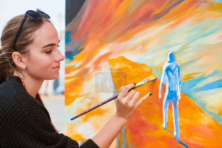 Attractive female painter passionately draws picture with paintbrush for outdoor street exhibition, close up side view of female artist infusing life into outdoor art space