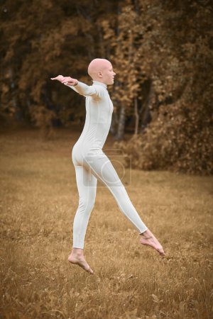 Téléchargez les photos : Portrait complet de jeune fille sans poils ballerine avec alopécie en costume blanc serré se prépare à sauter sur la pelouse d'automne dans le parc, symbolisant surmonter les défis et l'individualité d'acceptation gracieuse - en image libre de droit