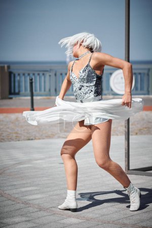 Joven chica sexy en el espacio micro falda de plata bailando con bufanda de seda blanca saludando con gracia, actuación de danza al aire libre femenina en el paseo marítimo creando un espectáculo al aire libre excitante