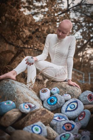 Young hairless girl with alopecia in white futuristic costume pensively examines stone with eye at surreal landscape, performance symbolizes introspection and reevaluation of individuality
