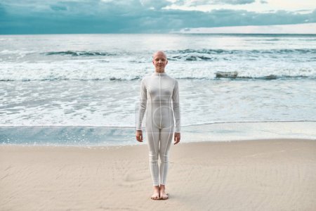 Ganztägiges Porträt eines jungen haarlosen Mädchens mit Alopezie im weißen futuristischen Anzug, das am Meeresstrand steht, metaphorische surreale Szene mit einem glatzköpfigen hübschen Teenager-Mädchen strahlt Zuversicht und einzigartige Schönheit aus