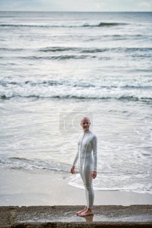 Junges haarloses Mädchen mit Alopezie im weißen futuristischen Anzug steht auf Betonzaun auf Meeresgrund, Glatze hübsches Teenager-Mädchen symbolisiert Mut und Akzeptanz der einzigartigen Erscheinung