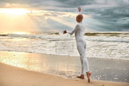 Junge haarlose Ballerina mit Alopezie im weißen futuristischen Anzug tanzt am Ufer des Meeres bei Sonnenuntergang, metaphorische surreale Performance eines glatzköpfigen hübschen Teenagermädchens strahlt Zuversicht, Hoffnung und einzigartige Schönheit aus