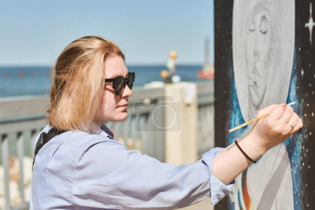 Young female painter in sunglasses passionately paints picture with paintbrush for outdoor street exhibition, female artist engrossed in creating vibrant artwork at bright sunny day