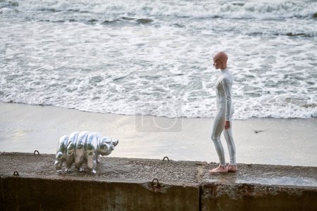 Joven chica sin pelo con alopecia en traje futurista blanco caminando sobre valla de hormigón con juguete tardígrado en el fondo del mar, chica bonita calva simboliza el coraje y la aceptación de la apariencia única