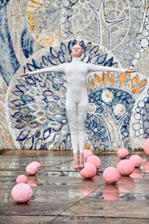 Junge haarlose Ballerina mit Alopezie im weißen futuristischen Anzug, die im Freien tanzt und zwischen rosa Kugeln auf abstraktem sowjetischen Mosaik springt, symbolisiert den Selbstausdruck
