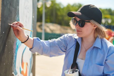 Junge Malerin mit schwarzer Mütze und Sonnenbrille malt leidenschaftlich Bild mit Pinsel für Straßenausstellung im Freien, Künstlerin in lebendige Kunstwerke bei strahlendem Sonnenschein vertieft
