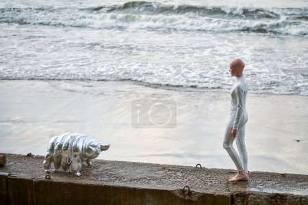 Jeune fille sans cheveux avec alopécie en costume futuriste blanc marchant sur une clôture en béton avec jouet tardif sur fond de mer, chauve jolie fille symbolise le courage et l'acceptation de l'apparence unique