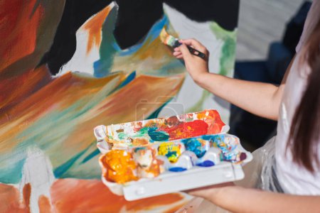Pintora mano apasionadamente pinta cuadro con pincel para exposición callejera al aire libre utilizando colores vibrantes, vista de cerca