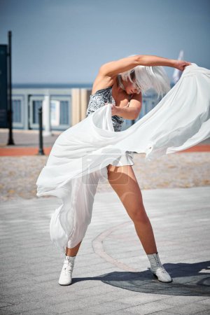 Foto de Joven chica sexy en el espacio micro falda de plata bailando con bufanda de seda blanca saludando con gracia, actuación de danza al aire libre femenina en el paseo marítimo creando un espectáculo al aire libre excitante - Imagen libre de derechos