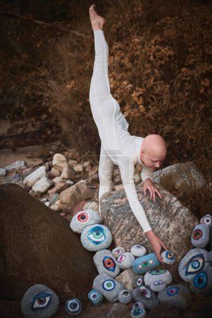 Flexible hairless girl with alopecia in white futuristic costume put her foot up and reaches hand for surreal landscape with lot of rocks with eyes, profound symbolism of embracing individuality