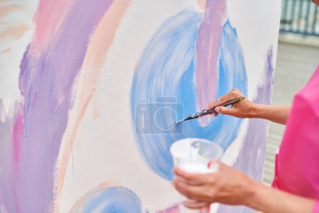 Female artist hand applies small brushstrokes of paint with paintbrush to canvas creating picture for exhibition, close up view of female hand painter creative process