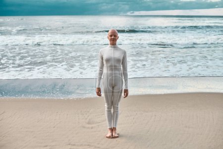 Ganztägiges Porträt eines jungen haarlosen Mädchens mit Alopezie im weißen futuristischen Anzug, das am Meeresstrand steht, metaphorische surreale Szene mit einem glatzköpfigen hübschen Teenager-Mädchen strahlt Zuversicht und einzigartige Schönheit aus
