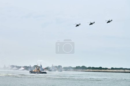 Photo for Fireboat sailing along Russian naval forces parade warships with military helicopters along coastline, seafaring tradition of military ships formation, nautical spectacle of russian sea power - Royalty Free Image
